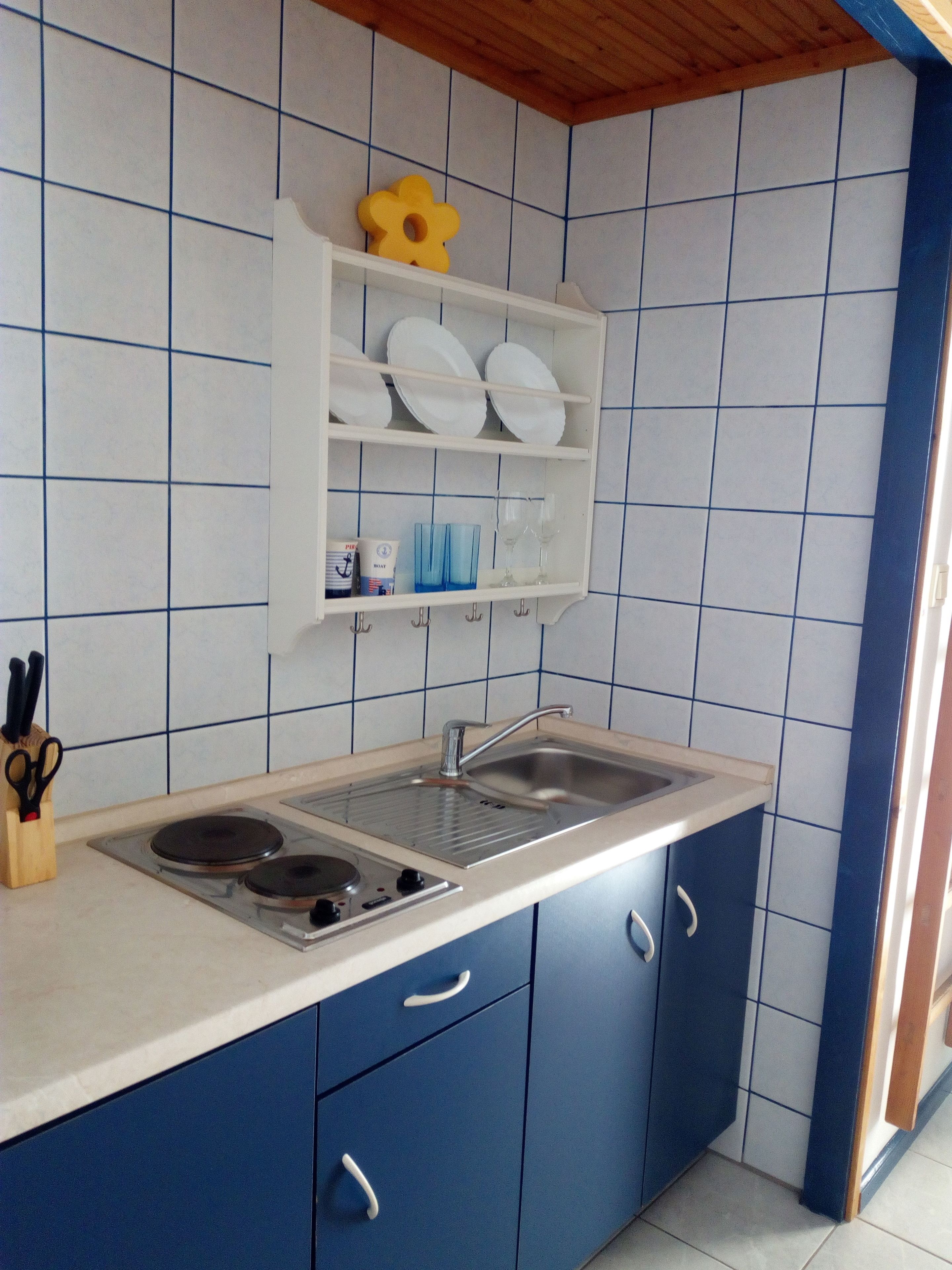 Una cocina con encimera blanca, armarios azules, un lavabo de acero inoxidable y una estantería blanca con vajilla y una flor amarilla.