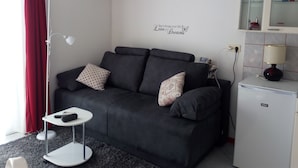 Un salon moderne avec un canapé noir, des coussins à motifs, une petite table d’appoint blanche, un lampadaire et un mini-réfrigérateur.
