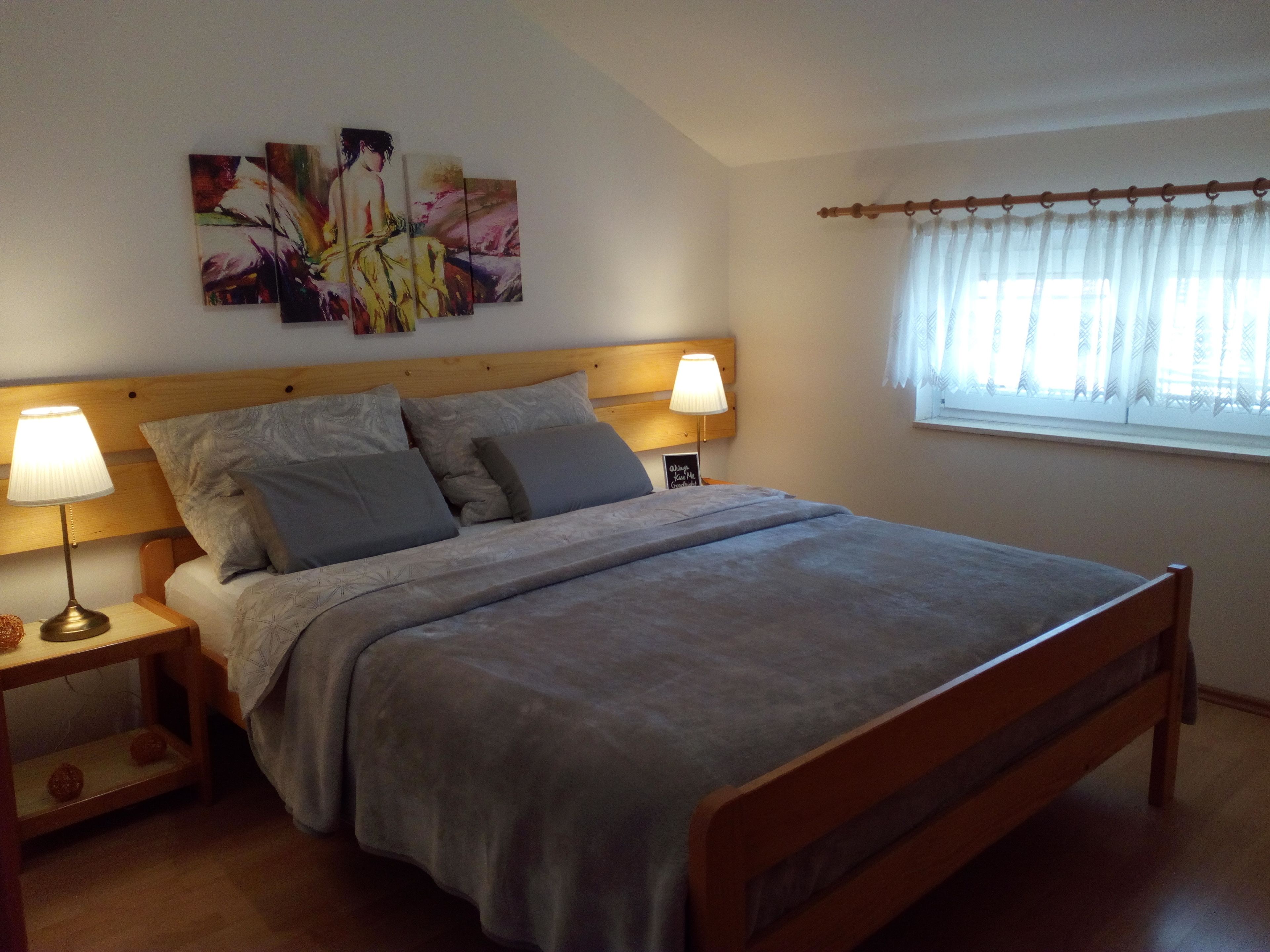 A bedroom with a bed, two bedside tables with lamps, a window with curtains, and a wall with a painting.
