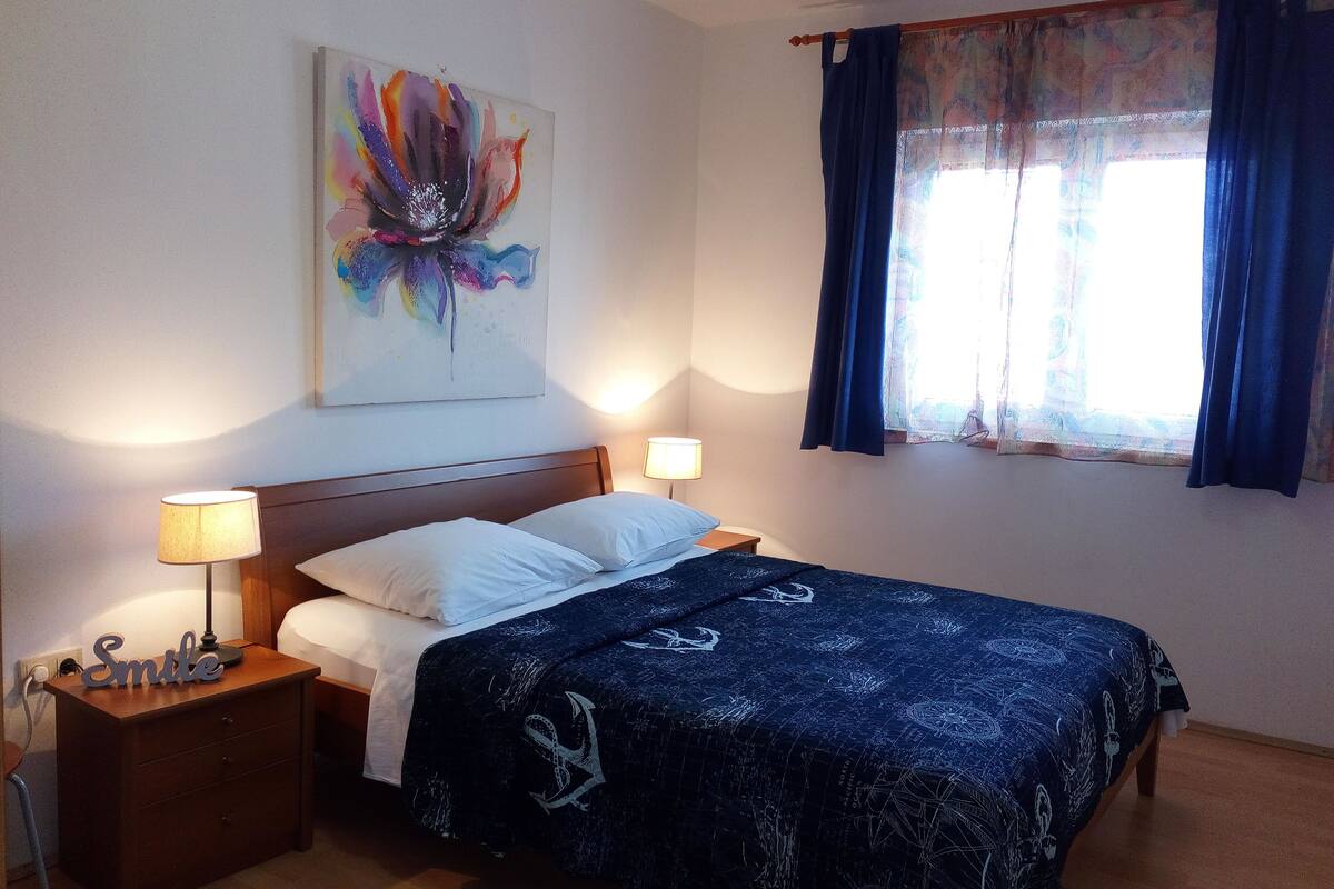 A bedroom with a bed, two bedside lamps, a nightstand, a wooden headboard, a wall-mounted painting, and a window with curtains.