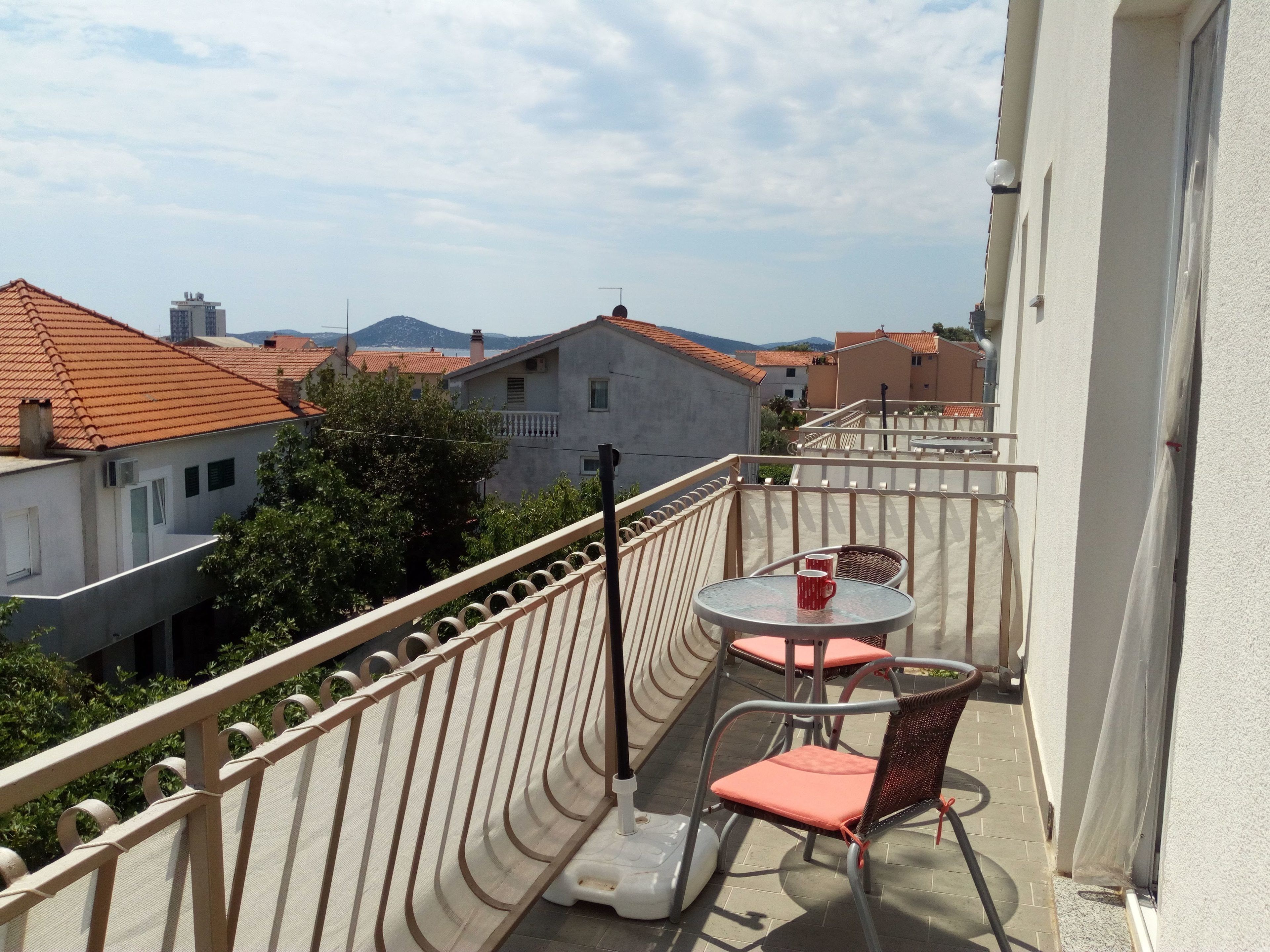 Un balcon avec une table et des chaises, donnant sur un quartier résidentiel avec des maisons aux toits rouges et une montagne visible au loin.