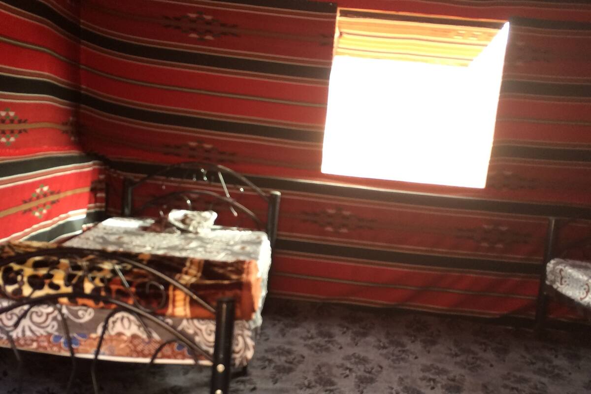 A room with a patterned carpet, a bed with a patterned bedspread, a small table with a chair, and a window with a view of the outside.
