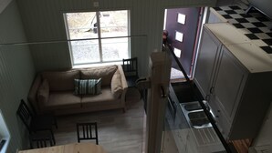 Habitació d'estil loft amb sostre alt, un sofà amb coixí escocès, una taula de dinar i una zona de cuina amb terra de pissos.