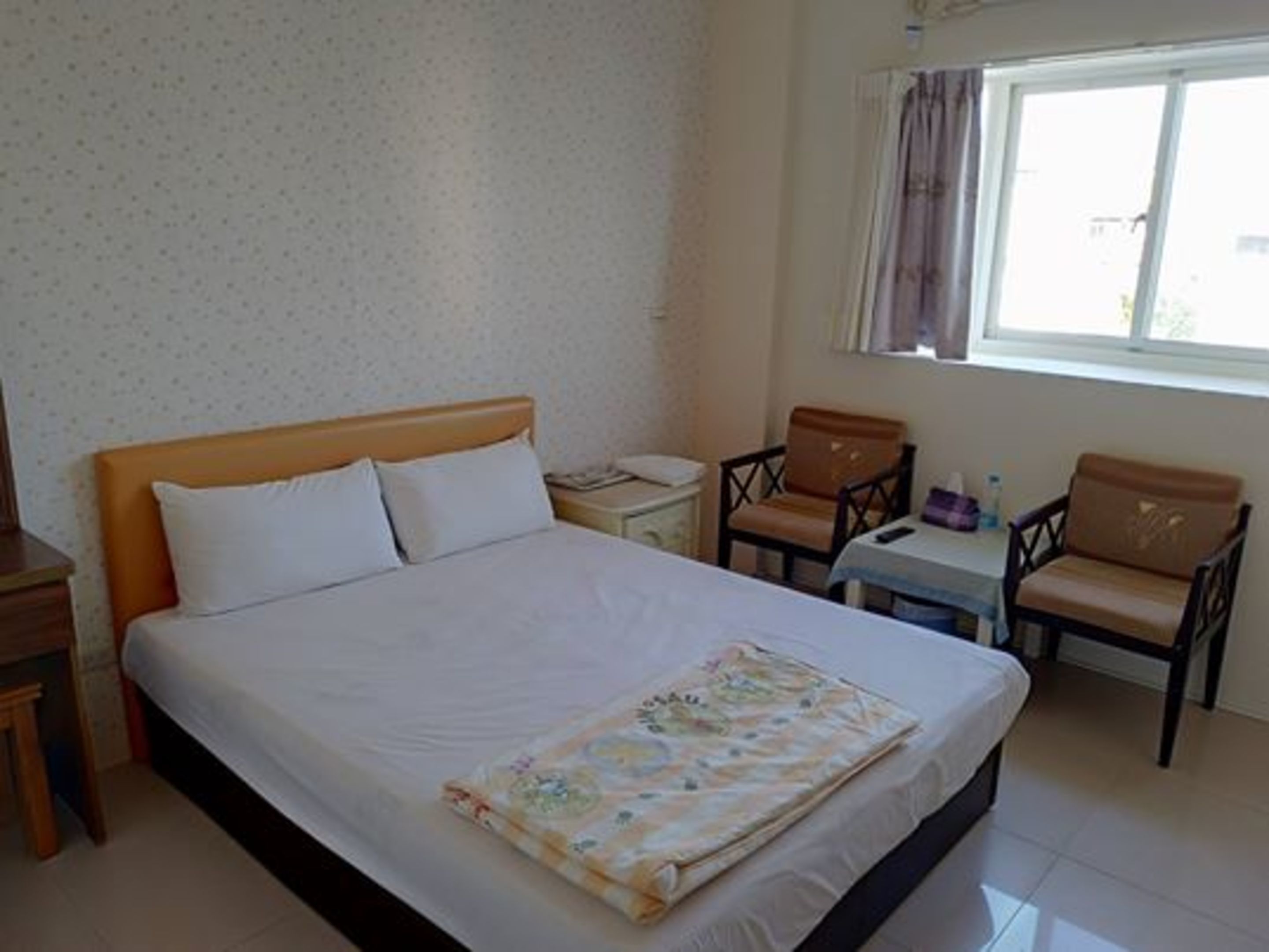 A hotel room with a bed, a small table with chairs, a window with curtains, and a wall with a subtle pattern.