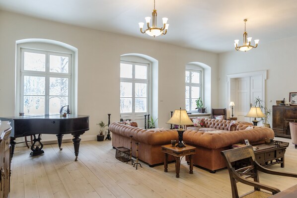 A spacious living room with a grand piano, a large brown sofa set, and elegant chandeliers.