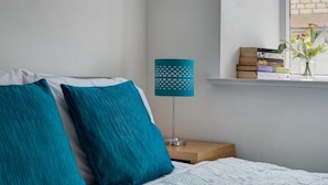 A neatly made bed with teal pillows, a bedside lamp, and a stack of books on a nightstand.