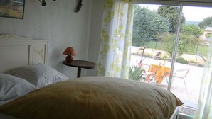 A bedroom with a large bed, a small table, and a view of the outdoors through a window. - B&B Chez Rose-Marie (Vaison-la-Romaine)
