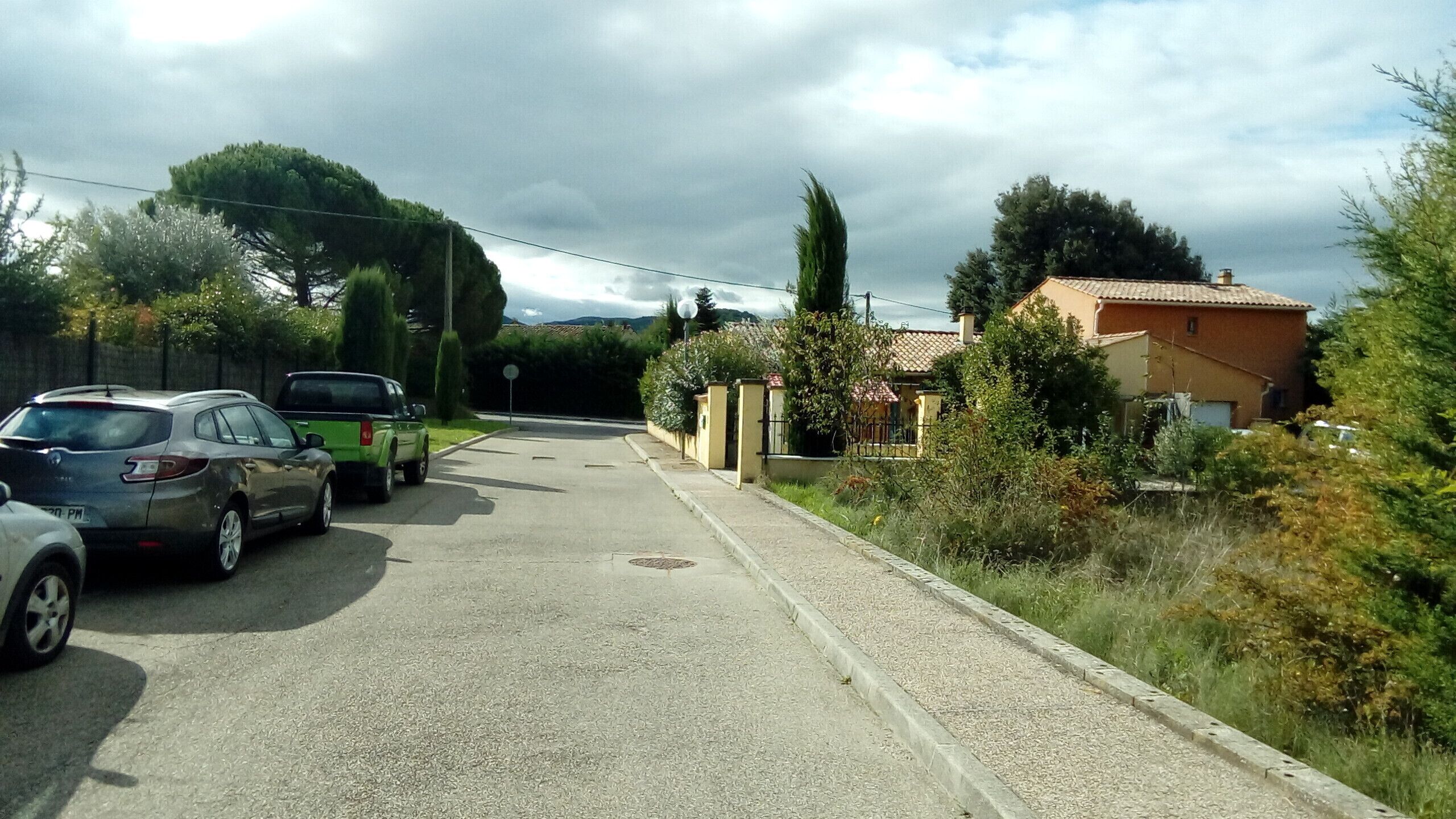 Uma rua residencial com carros estacionados, uma casa com telhado em tijolos e vegetação exuberante.
