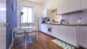 A modern kitchen with white cabinets, a microwave, an oven, and a sink. There is a window with blinds, a table with chairs, and a patterned rug on the floor. - Italianway Corso Lodi 121 (Milan)