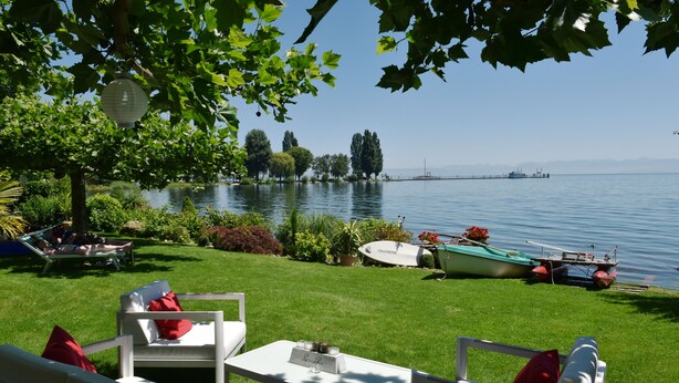 Un patio junto al lago con muebles de exterior blancos, una lancha amarrada cerca y una vista despejada del lago y montañas distantes.