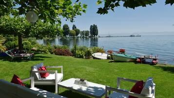 A lakeside patio with white outdoor furniture, a boat docked nearby, and a clear view of the lake and distant mountains.