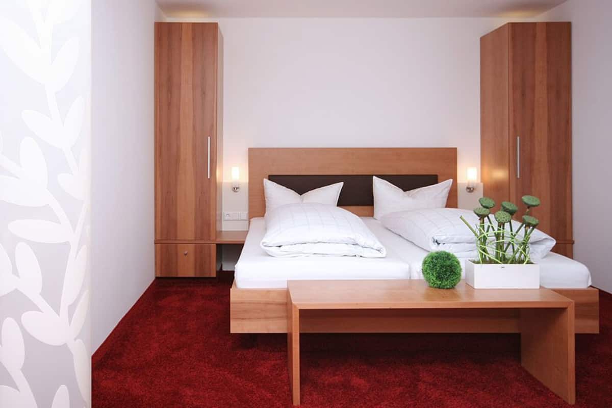 Un dormitorio con cabecera de madera, dos almohadas blancas, una cama bien tendida, una mesita de noche pequeña y una planta en maceta sobre la mesita.