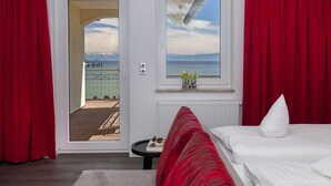 Habitación de hotel con cama, una mesita, ventana con vistas al mar y a las montañas, y cortinas rojas.