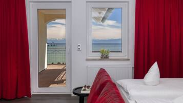 A hotel room with a bed, a small table, a window with a view of the sea and mountains, and red curtains.
