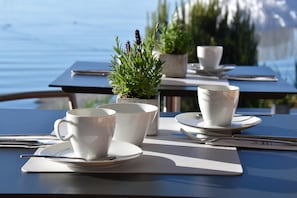 A table set with white cups, saucers, and cutlery, with a small potted plant and a view of the sea in the background. - Strandhaus Eberle (Immenstaad am Bodensee)