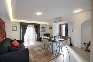 A modern living room with a sofa, dining table, and chairs. There is a window with curtains, a potted plant, and a wooden sculpture. - Apartment/ flat - Taormina (Taormina)