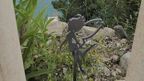 Une sculpture représentant une figure tenant une coupe, entourée de verdure et surplombant une étendue d’eau avec, au loin, une terre ferme.