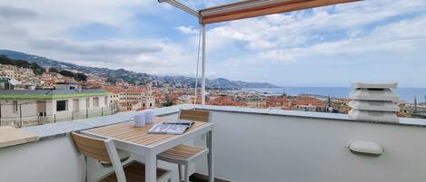 A balcony with a table and chairs, overlooking a coastal cityscape with buildings and a view of the sea.