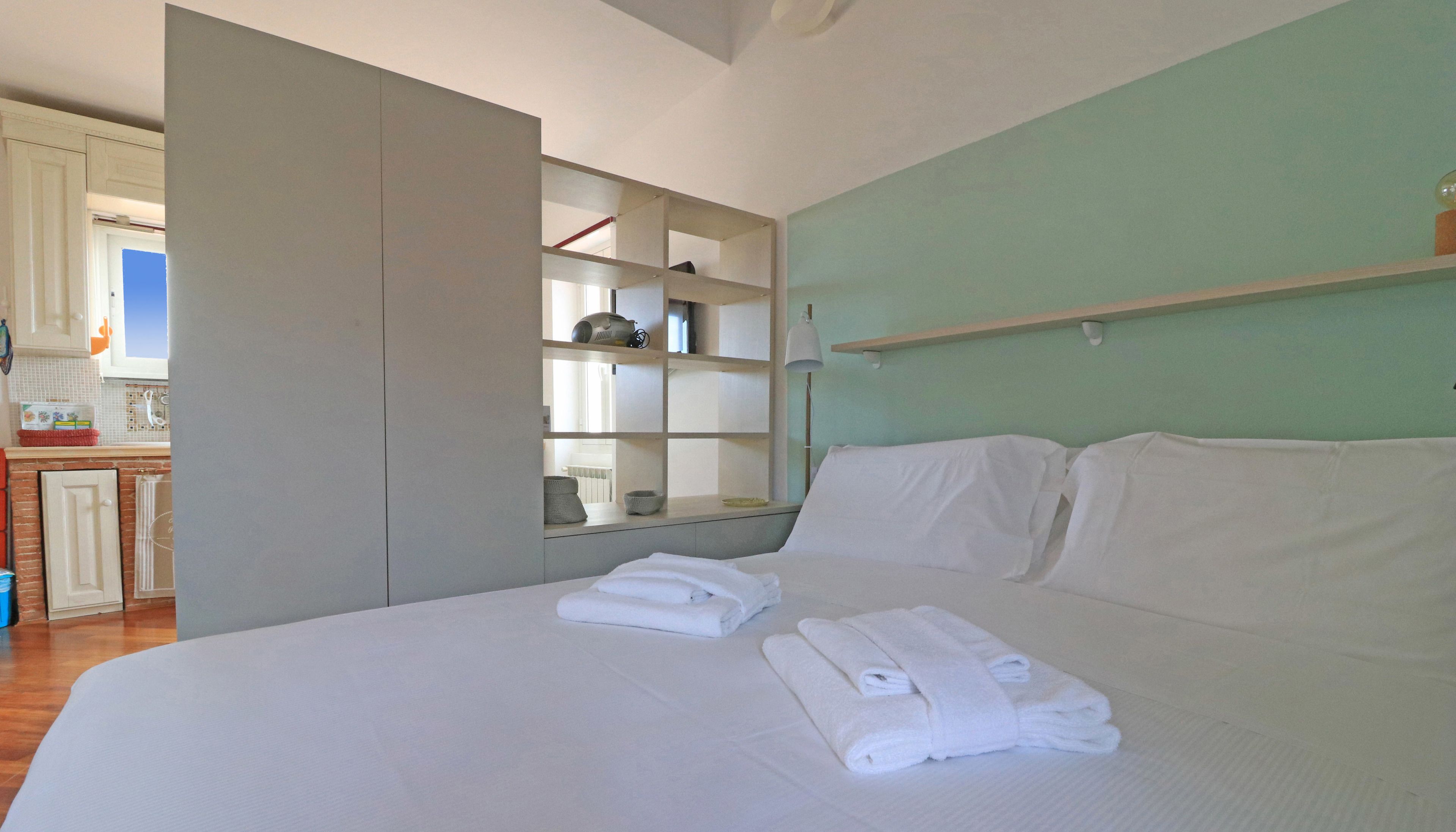 A bedroom with a bed, white towels, a shelf with various items, and a view of a kitchen area.