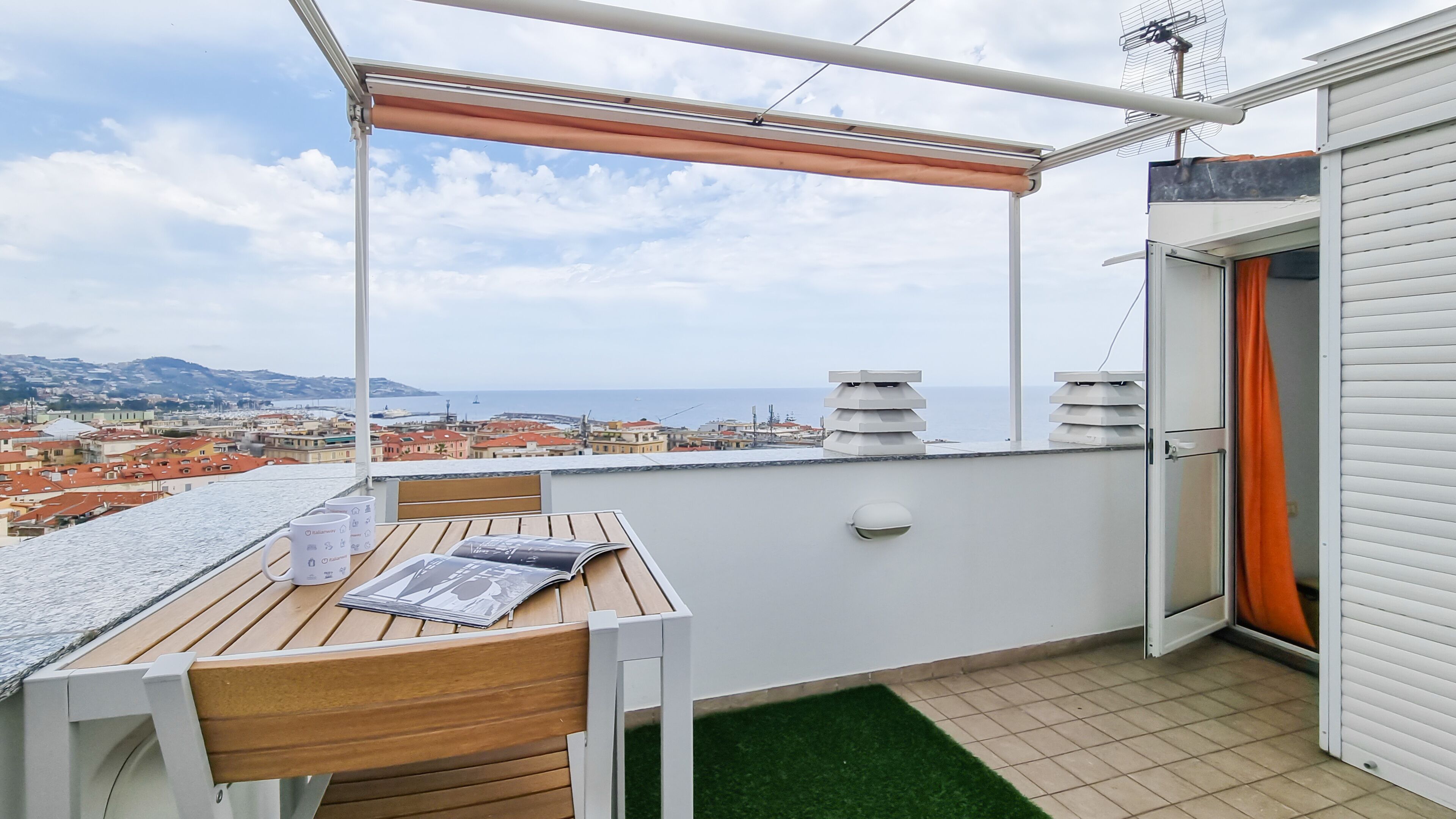 Un balcone con tavolo e sedie, vista sul mare e sulla città, e una porta con una tenda arancione.