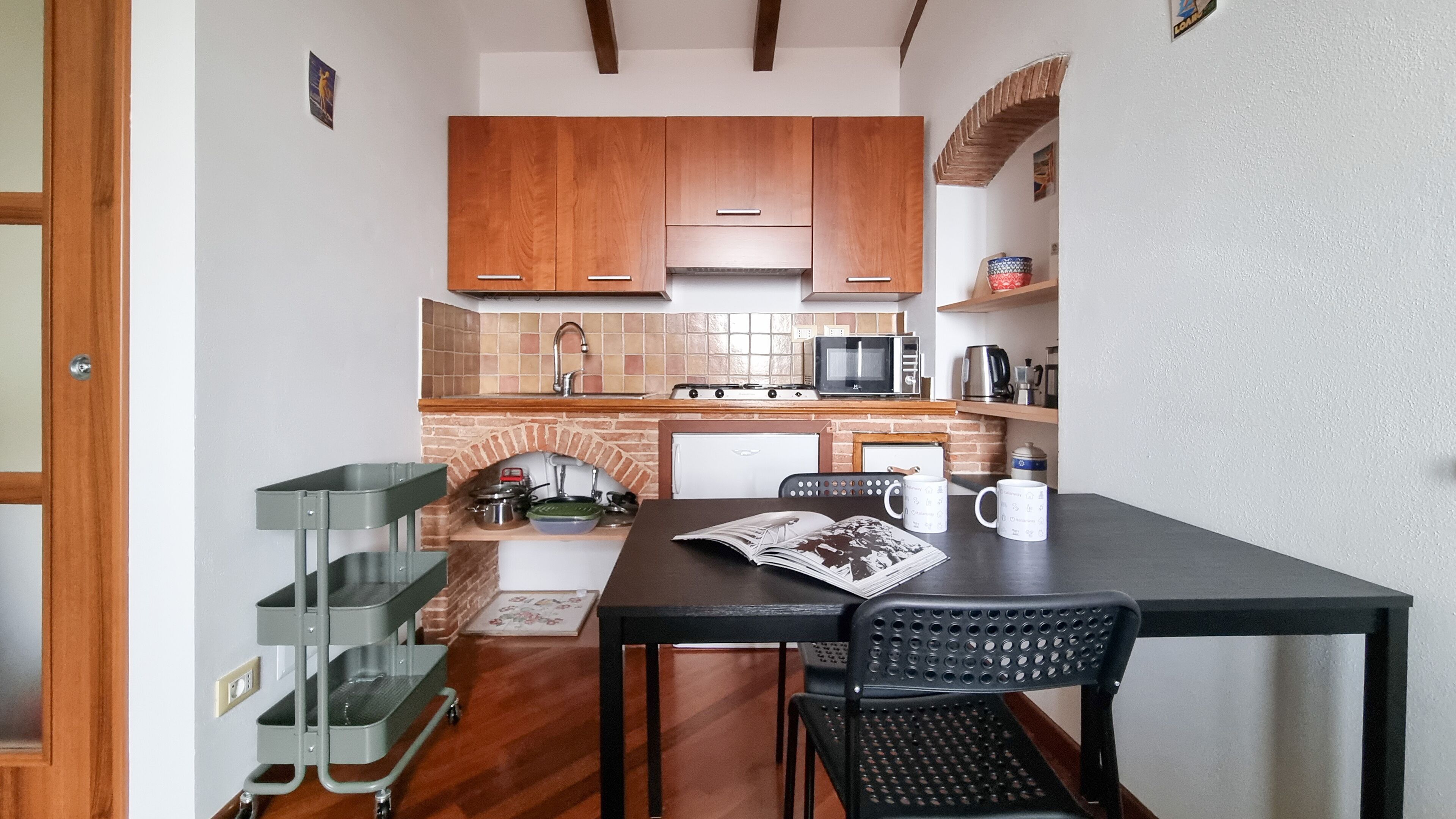 A compact kitchen with wooden cabinets, a microwave, a stove, and a coffee maker. There is a dining table with chairs and a magazine on it.