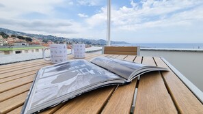 A wooden table with a magazine and two mugs with Italianway branding, overlooking a coastal town and sea. - Italianway Costiglioli (Sanremo)