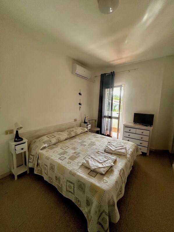 A bedroom with a bed, a TV, a bedside table, a lamp, and an air conditioning unit.