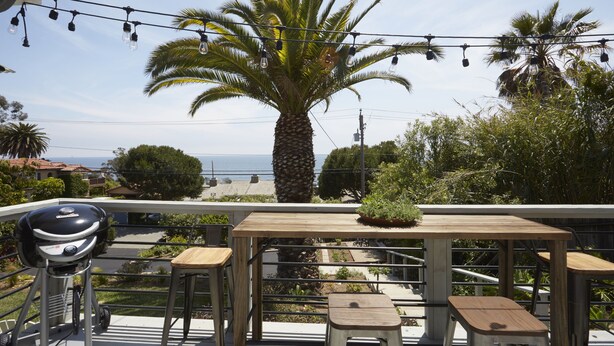 A balcony with a wooden table and stools, overlooking a beach and palm trees. - Surf Bungalow at the Lazy Hummingbird (Summerland)