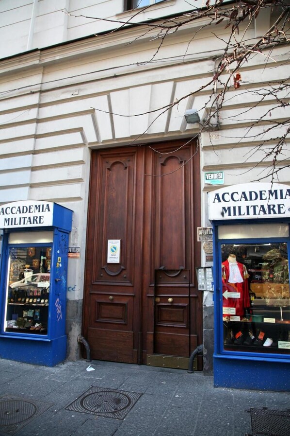 Fachada de edificio con un gran puerta de madera, dos escaparates azules con la señalización 'Accademia Militare' y un maniquí en el escaparate de la derecha.