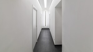 Un pasillo moderno y estrecho con paredes blancas y suelo oscuro, con iluminación empotrada y una puerta a la izquierda.