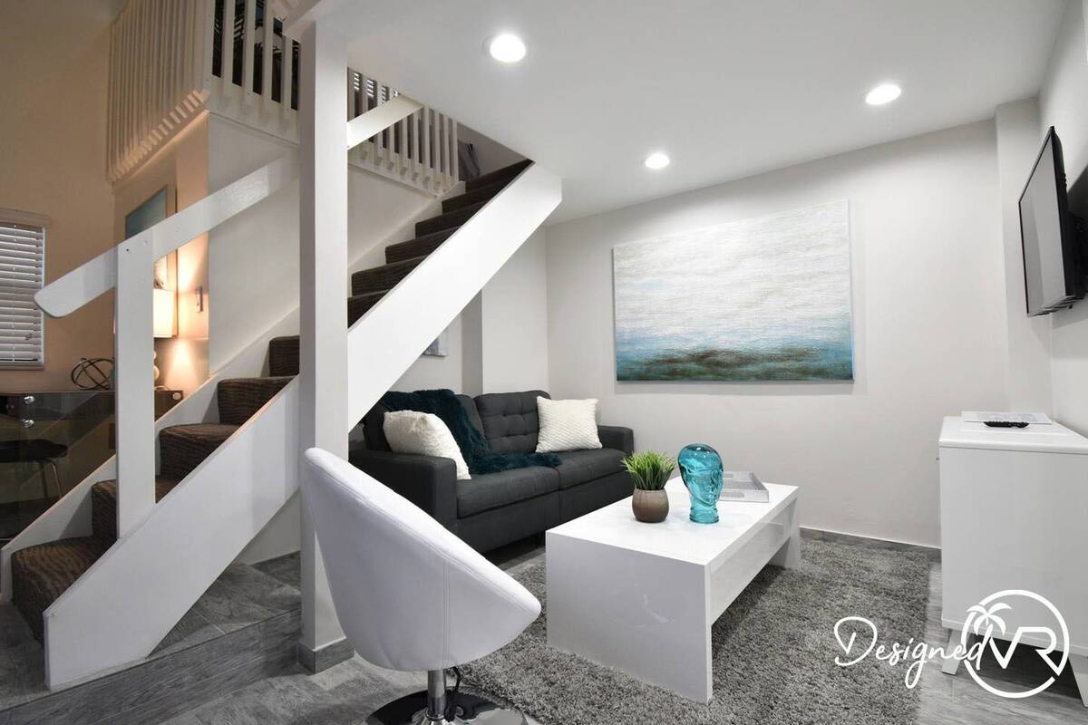 A modern living room with a staircase, a grey sofa, a white coffee table, and a painting on the wall.