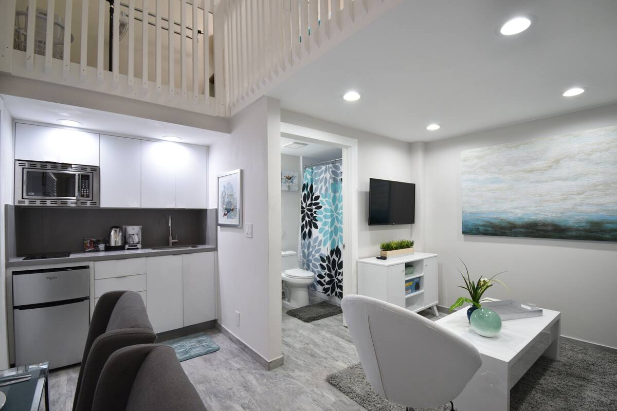 A modern kitchen with a microwave, refrigerator, and coffee maker. There is a dining area with a white table and chairs, and a TV mounted on the wall.