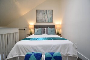 A bedroom with a bed, two bedside lamps, a wall-mounted painting, and a window with blinds. - Hobe Lofts (Hollywood)