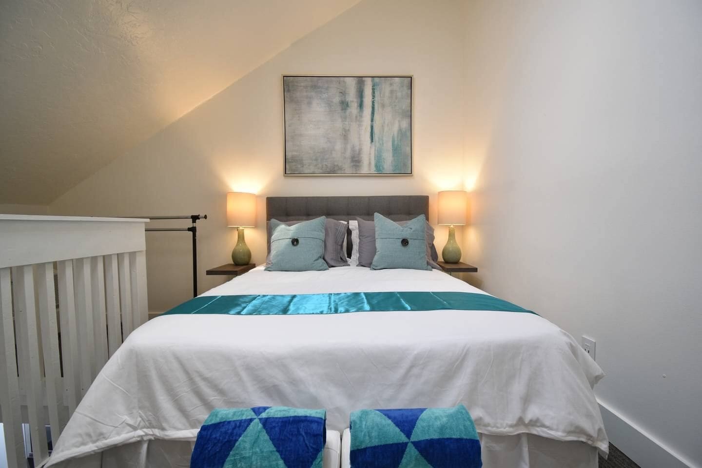 A neatly made bed with a white and teal bedspread, two matching pillows, and two bedside lamps on either side.