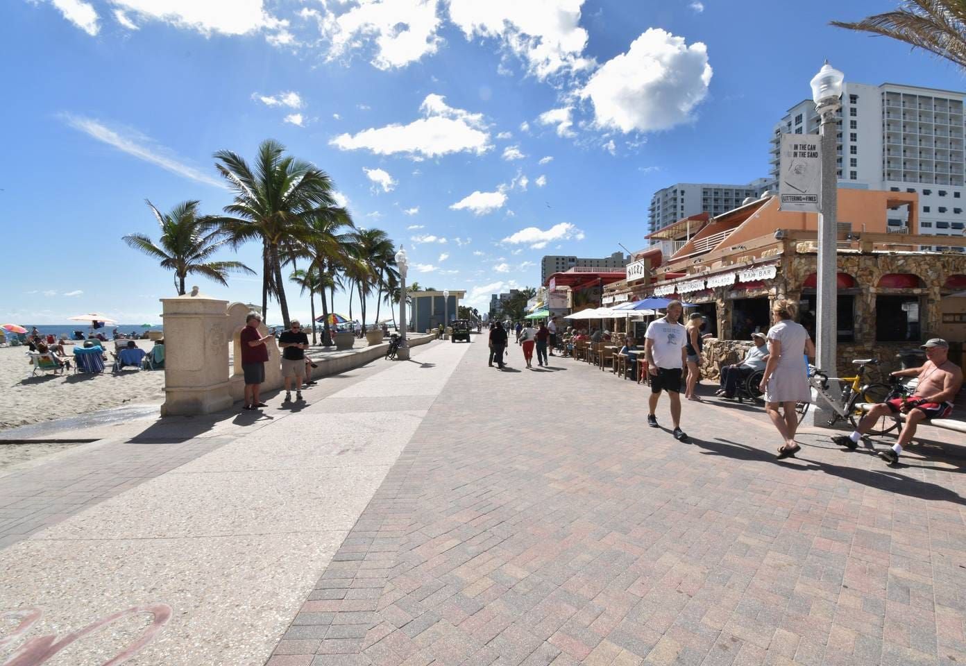 Une promenade dallée bordée de palmiers, de parasols de plage et où l’on peut voir des gens se promener.