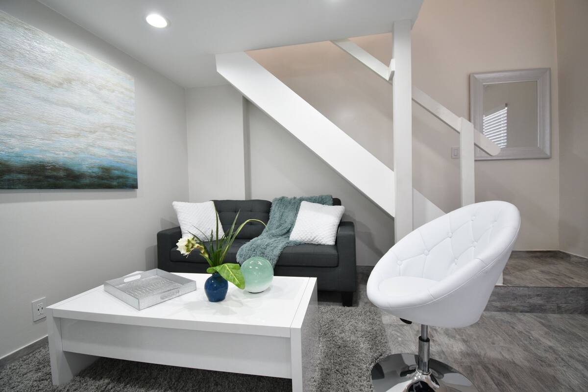 A modern living room with a white coffee table, a black sofa with a blue throw, a white chair, and a staircase.