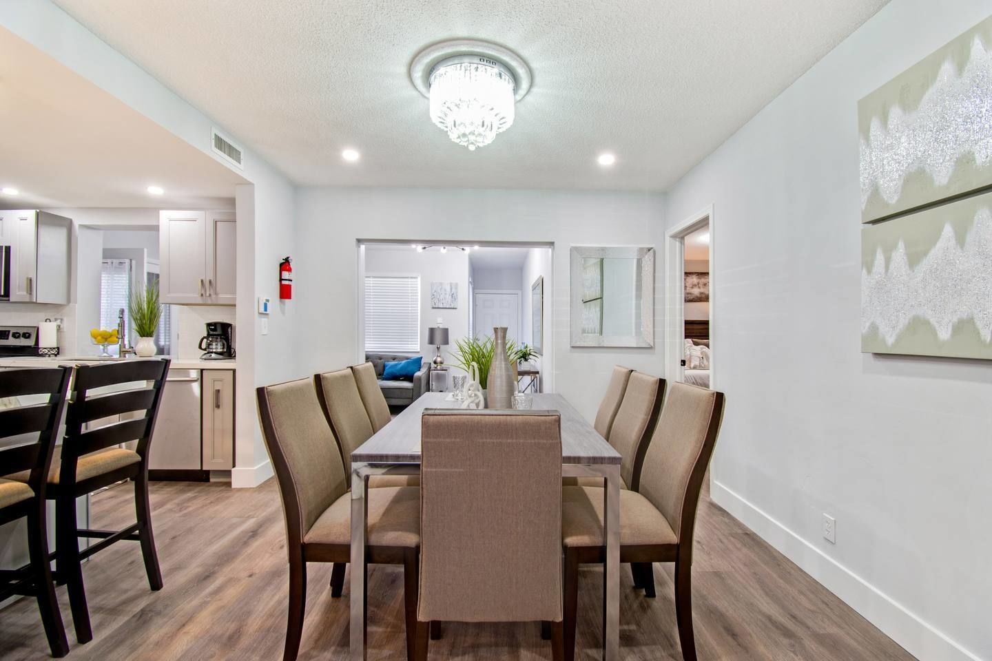 A modern dining area with a long table and six chairs, a chandelier, and a kitchen in the background.