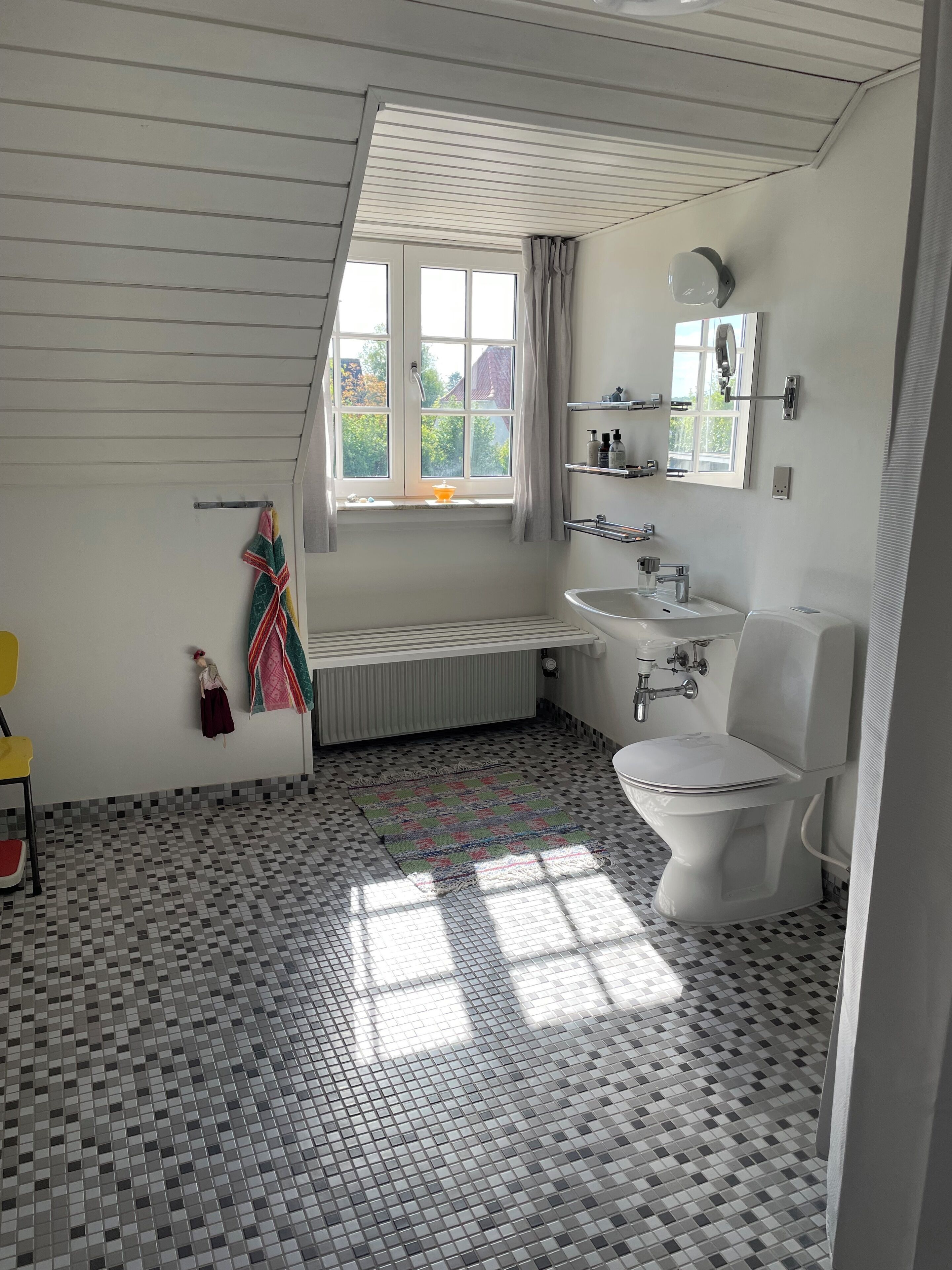 Un baño con piso a cuadros, una ventana con cortinas, un radiador, un lavamanos, un inodoro y un perchero para toallas.