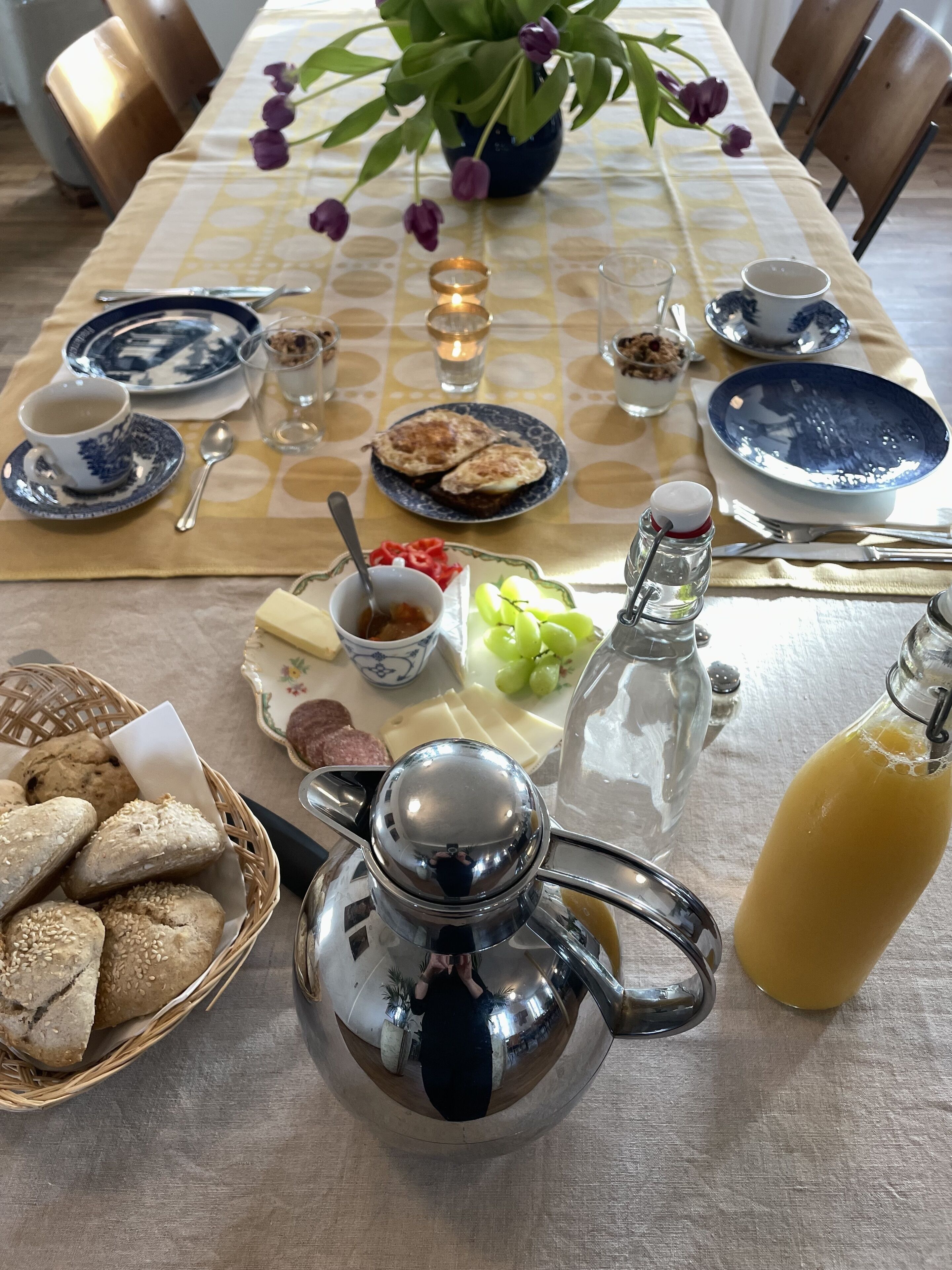A breakfast setting with a table set for two, including bread, cheese, fruit, and a coffee pot.