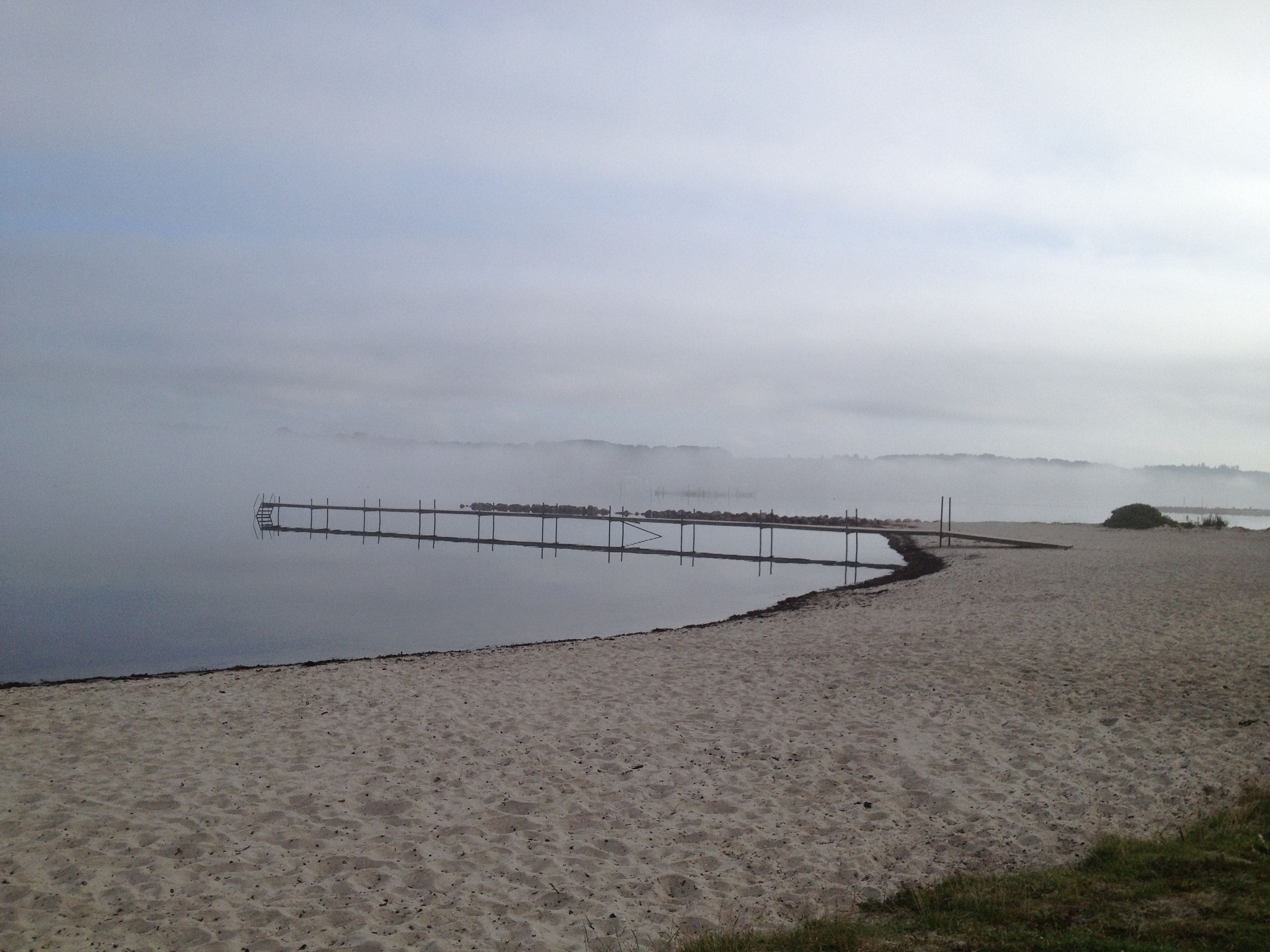 A beach with a wooden pier extending into calm waters, surrounded by foggy terrain.