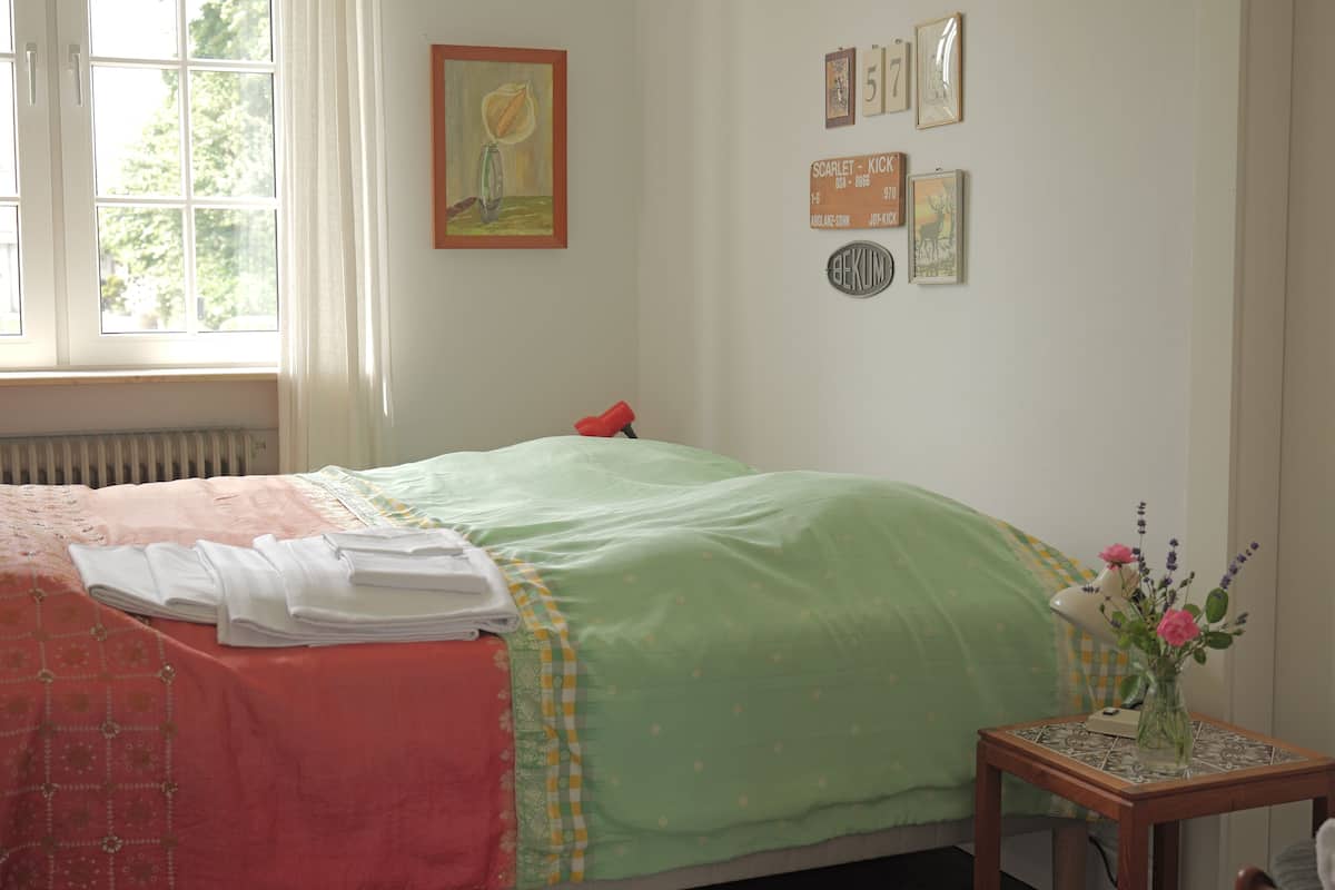 Un dormitorio con cama, radiador, ventana, mesita de noche con un florero y adornos en la pared.