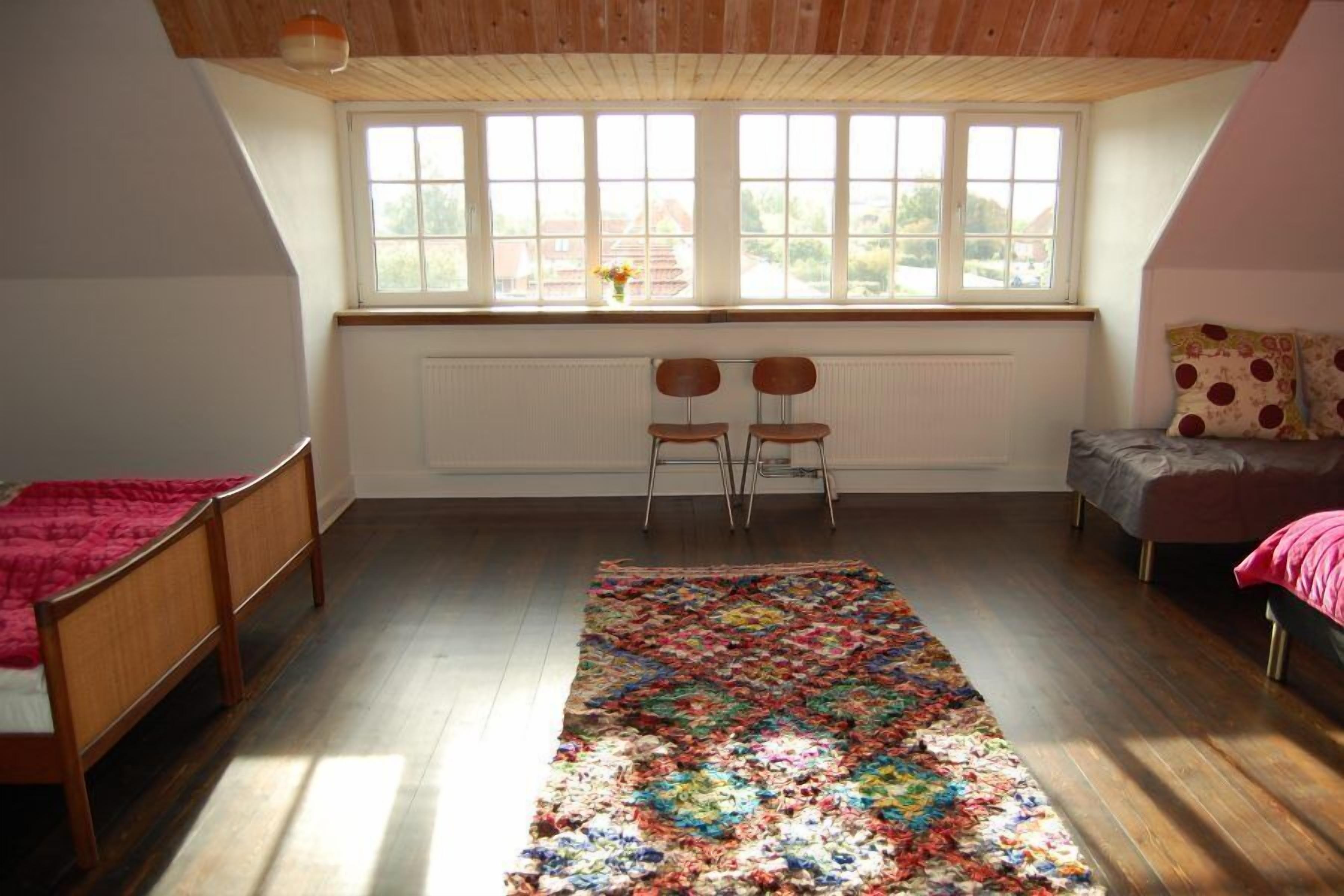 Une pièce avec un plafond en pente, un plancher en bois, une grande fenêtre, un lit, une chaise, une petite table et un tapis coloré.