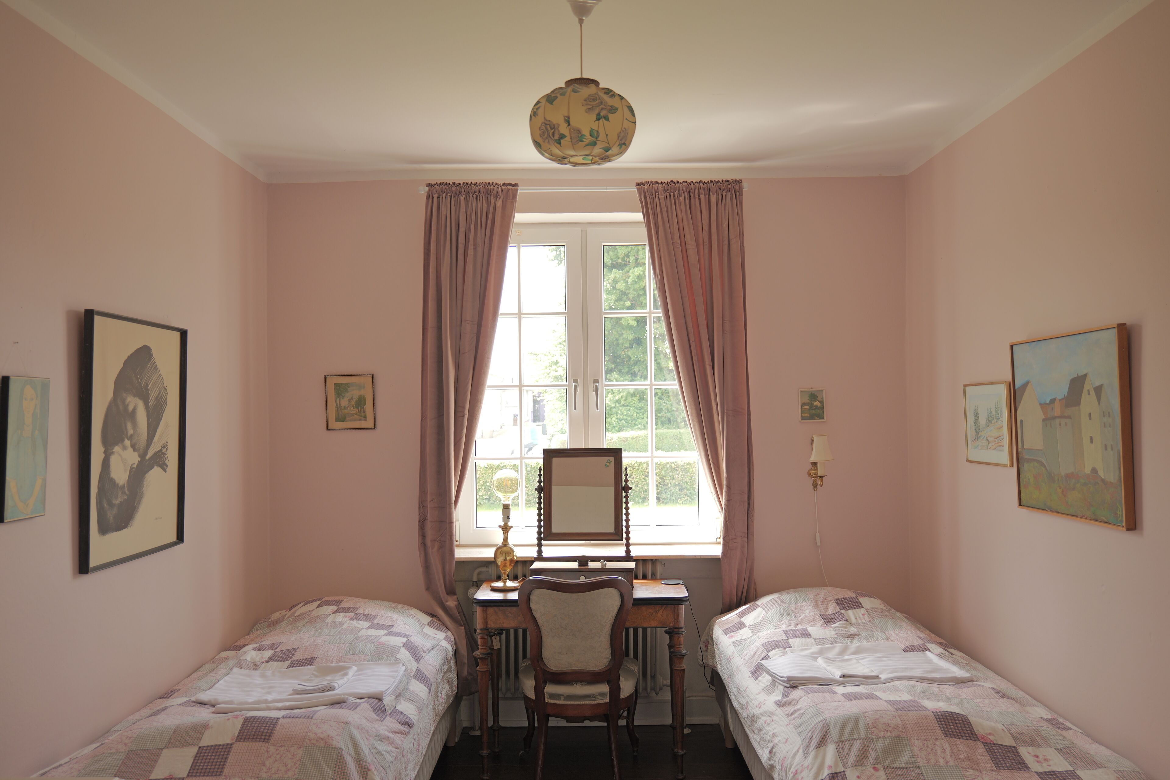 A bedroom with two beds, a desk, and a chair. There are curtains on the window, and artwork on the walls.