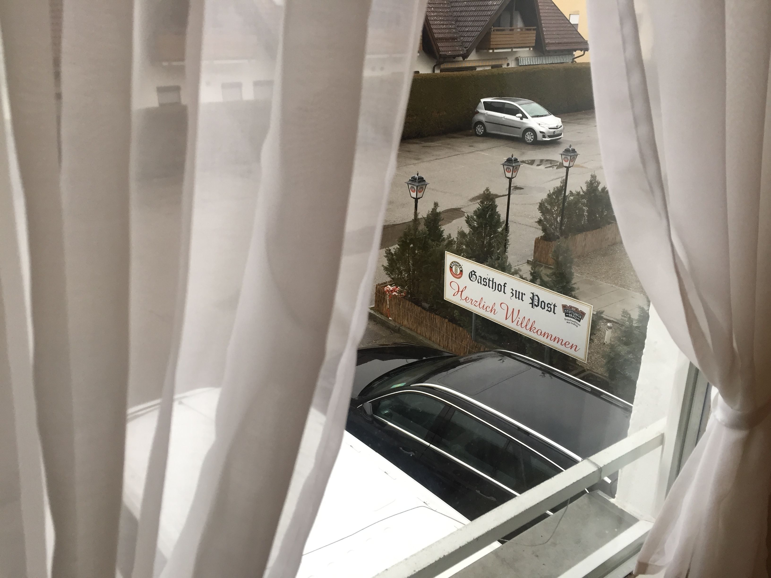 A view through a window with curtains, featuring a sign that reads "Gasthof zur Post" and "Herzlich Willkommen" with a logo.