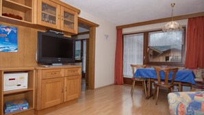 A compact living space with a wooden cabinet, a television, a dining area with a table and chairs, and a window with curtains.