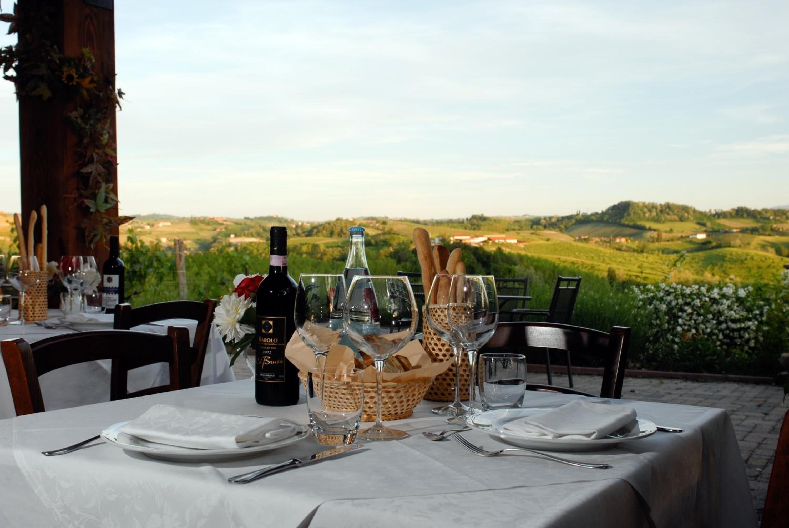 Mesa de jantar ao ar livre posta com copos de vinho, pratos e uma garrafa de vinho, com vista para uma paisagem deslumbrante.