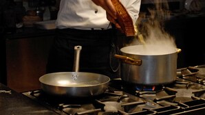 Een kok in een witte uniform en koksmuts kookt in een keuken met een pan op het fornuis en een koekenpan op het aanrecht.