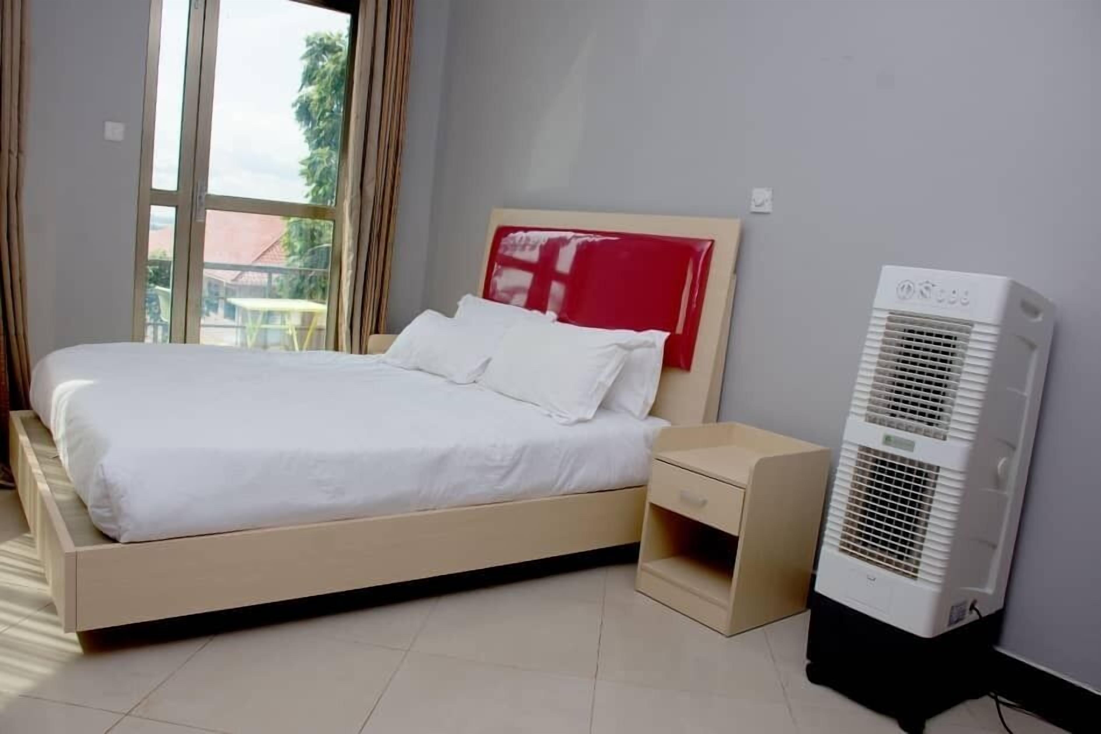 A neatly made bed with white linens, a bedside table with a drawer, and a standing air conditioner.