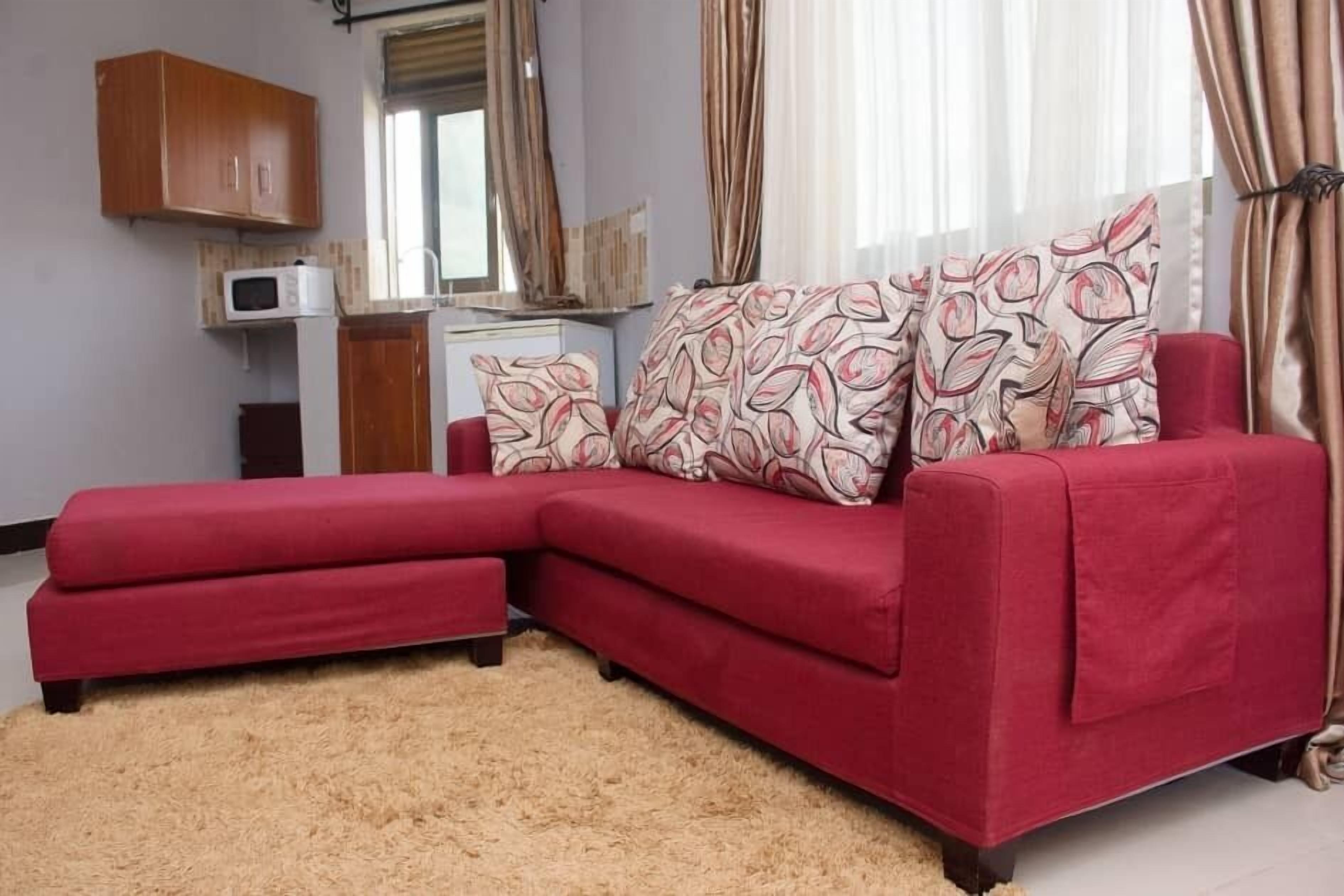 A red sectional sofa with patterned cushions, a small kitchenette with a microwave, and a window with curtains.