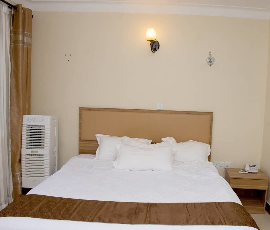 A hotel room with a bed, a bedside table with a phone, a wall-mounted light, and an air conditioning unit.